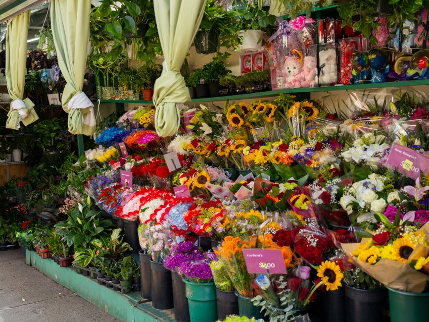 An outdoor flower display at a florist shop featuring a variety of fresh floral bouquets and arrangements. The assortment includes vibrant sunflowers, roses, lilies, and other seasonal flowers wrapped