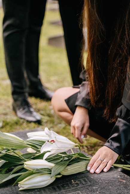 A person kneels on the ground, arranging a bouquet of white lilies with long green leaves on a dark stone memorial at a funeral site. The lilies have large, open petals and red stamens, and are wrappe