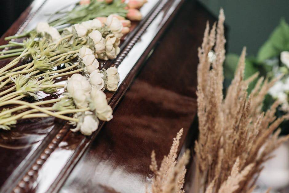 A close-up of a floral arrangement featuring a bouquet of fresh white spray roses with delicate petals and green stems, placed on a polished dark wooden surface. The bouquet is wrapped in clear cellop