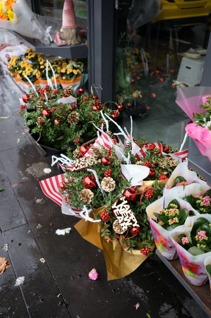 A festive floral arrangement displayed outside a flower shop showing a bouquet of fresh greenery, red berries, pinecones, and small white flowers, all wrapped in brown paper with a red and white strip