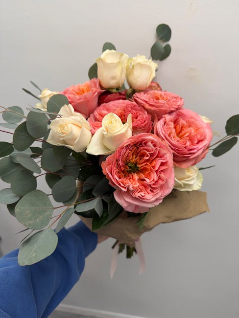 A bouquet of fresh flowers held by a person in a blue sleeve, featuring large, ruffled pink and coral ranunculus blooms, creamy white roses, and sprigs of eucalyptus with broad, rounded green leaves, 