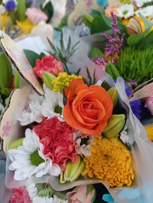 A vibrant bouquet featuring an orange rose as the focal point, surrounded by white daisies, pink carnations, yellow chrysanthemums, and purple statice flowers, all arranged with green foliage. The flo
