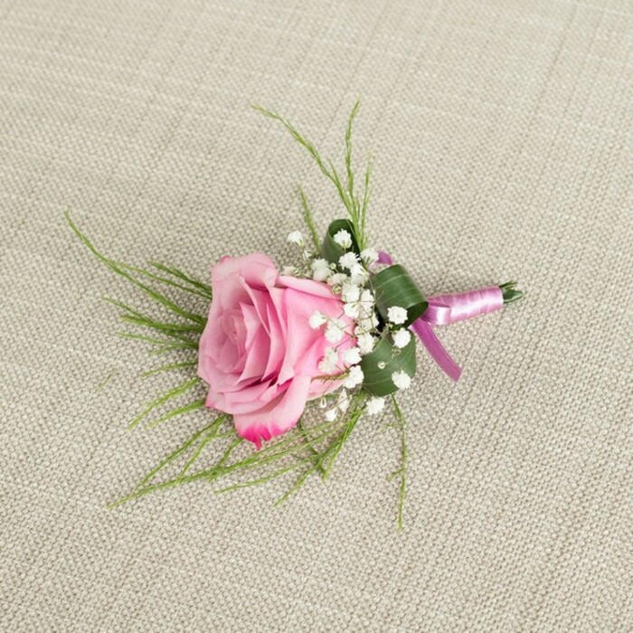 A finely crafted boutonniere featuring a single blush pink rose is displayed on a neutral, textured beige linen cloth, the kind of calm, natural backdrop you might see in a South Croydon bridal suite as final touches are added before the ceremony. The photograph is taken from directly above, capturing the full shape of the design in clear detail. The rose sits at the centre, semi-open and velvety, its petals spiralling out from the tight heart of the bloom. Near the centre, the petals are a soft, delicate pink that gradually intensifies to a deeper, more romantic tone at the curled outer edges, hinting at both youthfulness and maturity in one flower. Surrounding the base of the rose are clusters of white baby's breath, each tiny blossom forming part of an airy, cloud-like ring that softens the transition between the central flower and the foliage. Wispy strands of vivid green, grass-like foliage extend from behind the rose, fanning outward in graceful arcs and adding a sense of lightness and movement. Nestled beneath, a single darker green leaf is carefully folded and wrapped around the stems, giving the piece a clean, tailored finish. The stems are bound in a smooth satin ribbon that shifts between soft lavender-pink and a richer magenta-purple, the subtle sheen of the fabric catching the light and echoing the rose's colour. The ribbon is tied securely, with a compact knot and gently tapered ends. A small, metallic silver pin peeks out near the base, ready to fasten the boutonniere securely to a lapel. The overall mood of the image suggests quiet elegance and romantic intention, ideal for weddings, proms, or black-tie events around South Croydon, from intimate gatherings near Croham Hurst to celebrations along the quieter residential streets nearby.