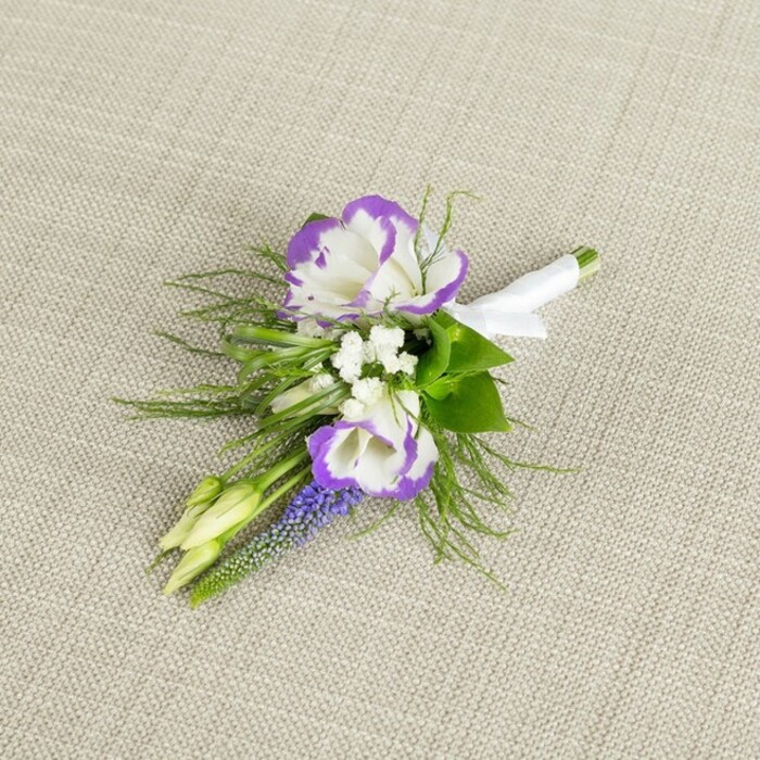 On a softly textured, light beige linen background, a single elegant boutonniere has been carefully placed, the neutral surface acting like a canvas that lets the colours and textures of the flowers come alive. At its heart are two lisianthus blooms, fully open yet still delicate, their creamy white petals gently ruffled and edged with a rich, velvety purple that forms a striking halo around each flower. Close by, tiny white filler flowers-resembling fine sprigs of baby's breath-cluster in small, cloudlike tufts, easing the transition between the bold two-tone lisianthus and the deeper hues. Standing proudly among them, a slender spike of muscari, or grape hyacinth, draws the eye with its tightly packed, bell-shaped florets in a deep purple-blue, adding vertical interest and a slightly whimsical texture, as though freshly picked from a shaded garden corner near Haling Grove. Surrounding the central flowers, feathery greenery, much like asparagus fern, fans out in light, airy sprays, interwoven with a few broader, richer green leaves that lend structure and contrast. Two pale green buds are nestled within the foliage, their smooth surfaces and closed petals hinting at blossoms yet to open-perfect symbolism for a wedding morning or prom night. The stems are neatly gathered and aligned, expertly bound with a band of crisp white satin ribbon that wraps smoothly around the base, finished with a small, precise bow that lies flat and refined. This considered composition, with its balance of white, purple, and fresh green, feels both classic and contemporary, the sort of boutonniere a South Croydon groom might wear as he leaves his home near Brighton Road, stepping into a day filled with quiet anticipation and heartfelt celebration.