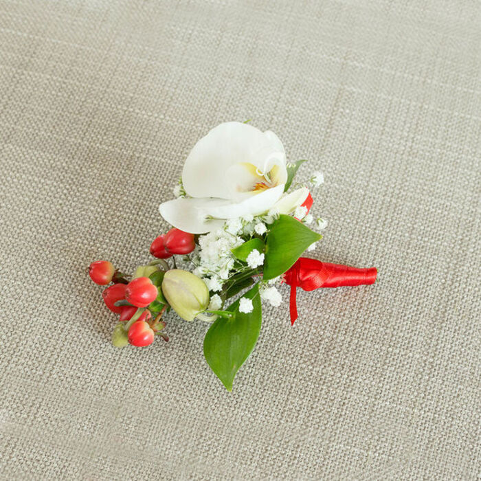 On a softly woven, light beige fabric that resembles natural linen or burlap, a carefully composed floral boutonniere is positioned as if waiting to be pinned before a ceremony in South Croydon. The central feature is a white Phalaenopsis orchid, its generous petals smooth and slightly curved, catching the light with a gentle, satin-like sheen. At the core of the orchid, delicate tones of creamy yellow and soft orange glow subtly, drawing the eye inward and adding warmth to the otherwise cool white bloom. Surrounding this focal flower, sprays of baby's breath form a light, cloud-like ring, each tiny white blossom poised on fine green stems that lend a whisper of texture and airiness to the design. On one side of the arrangement, a cluster of round, glossy red berries sits tightly together, their cherry-red skins reflecting a touch of shine, while a few paler green, under-ripe berries peep through, hinting at the natural progression of the seasons. Rich green foliage, ranging from slightly broader, pointed leaves to smaller, elongated ones, is woven between the flowers and berries, framing the composition with a fresh, botanical backdrop that makes every detail stand out clearly against the neutral cloth. The stems are gathered and wrapped in a vibrant red satin ribbon, carefully wound and secured in a neat knot, its short ends lying smoothly along the base and suggesting readiness for a lapel on a groom's jacket for a wedding near Sanderstead Road or a festive formal event by South Croydon station. The overall impression is one of classic elegance with a seasonal touch, combining refined orchid beauty with the joyful pop of berries for memorable local celebrations.