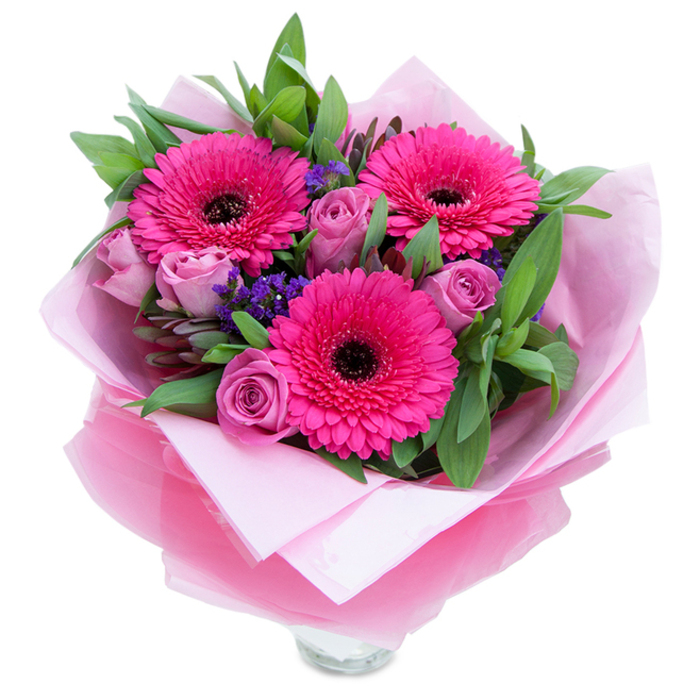 I see this arrangement as a polished embrace rendered in pinks and purples: three vivid fuchsia gerbera daisies dominate the scene, their radiating petals and pronounced dark centres giving the bouquet a bold, joyful heartbeat. Complementing them are several soft lavender-pink roses, their petals tightly rolled and gradually unfurling in a way that reads as both tender and composed. Interspersed statice sprigs in deep purple-blue add delicate points of texture, like wildflower confetti, while generous green foliage-suggestive of salal or eucalyptus-forms a lush backdrop that enhances the pink tones and roots the composition in natural freshness. The blooms are cocooned in an inner sheet of soft, matte pale pink tissue paper and an outer veil of translucent, slightly crinkled fuchsia-pink cellophane that reflects light with a gentle shimmer; the wrapping is folded with deliberate care and presented so the bouquet feels gift-ready. A clear glass vase rim shows at the base, indicating the florist has prepared the flowers for immediate display, perfect for a mantlepiece or kitchen table. As someone who arranges flowers for homes across South Croydon and nearby Sanderstead, I imagine this piece bringing quiet celebration to a mother on her special day or adding a romantic note to an anniversary dinner. The textures-the velvety rose petals, the crisp greenery, the papery statice-combine with a light, inviting scent to make this bouquet both comforting and exuberant.