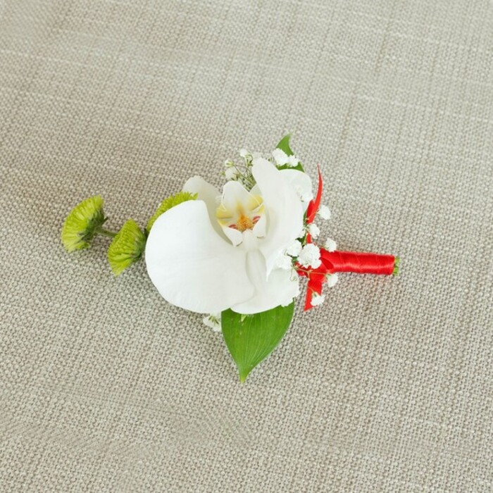 A sophisticated floral boutonniere lies horizontally on a light beige, linen-textured surface, the natural fibres of the fabric giving a relaxed, tactile backdrop that lets every detail of the arrangement stand out clearly, as if laid on a carefully prepared dressing table before a South Croydon ceremony. At its centre, a single white phalaenopsis orchid opens generously, each petal wide and smooth, with a gentle curve that suggests softness to the touch. The orchid's throat reveals a wash of soft yellow merging gracefully into fine reddish-orange or tawny markings, drawing attention to the intricate inner structure. Encircling the blossom, dainty sprays of white baby's breath cluster like tiny stars, their many small blooms adding an ethereal, cloud-like volume without overpowering the main flower. To the left of the orchid, two rounded lime-green chrysanthemum buds, dense with tightly packed, spiky petals, introduce a striking pop of contemporary green, reminiscent of fresh foliage in local parks and gardens around South Croydon. Supporting the composition underneath, a wide, deep green leaf provides a visual base, with a smaller, darker leaf visible closer to the stem, reinforcing the natural, layered look. The stems are expertly gathered and wrapped with a vivid crimson red satin ribbon, the fabric wound smoothly and securely, ending in a neat knot or tiny bow beneath the blooms. This bold red element adds a lively contrast to the whites and greens, echoing the celebratory energy of a wedding reception or formal event nearby. The lighting is soft and even, casting minimal shadows, which allows the glossy orchid petals, the matte baby's breath, and the satiny ribbon to each show their distinct textures. The boutonniere feels timeless yet modern, a handcrafted detail that would suit a groom, groomsman, or guest attending a special occasion anywhere in the South Croydon area, from intimate registry office vows to grand hotel celebrations.