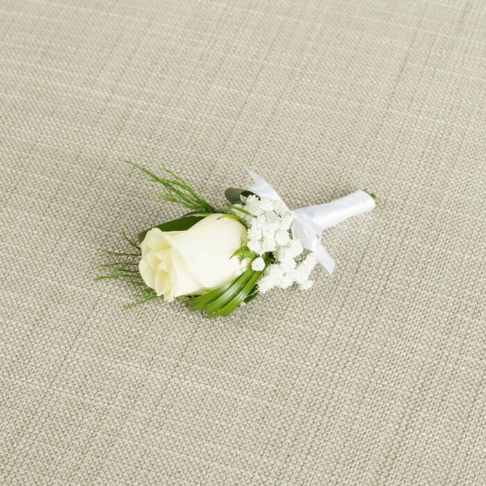 A single, meticulously made white rose boutonniere is shown resting at a graceful diagonal on a light beige, linen-like fabric, whose fine weave and gentle crosshatch pattern lend a natural, relaxed elegance to the scene. The focal point is a creamy white rosebud, medium in size and tightly furled, the outer petals wrapping smoothly around the inner ones to create a rounded, sculpted form that suggests both freshness and restraint. At the base of the bloom, delicate green sepals peek through, reinforcing the impression of a rose that has been carefully chosen and prepared shortly before the event, perhaps at a South Croydon florist serving couples celebrating near Selsdon Road or across to Haling Grove. Tucked to one side and partially cradling the rose are small, cloud-like clusters of white baby's breath; their many tiny blossoms give the boutonniere a light, airy quality and subtly enhance the rose's creamy tone. Behind and around the flowers, narrow, pointed leaves in a rich, deep green extend outward, some smooth and glossy, others more feathery and fern-like, offering variation in texture while maintaining a balanced, compact outline ideal for pinning to a lapel. The stems are gathered neatly and wrapped in a length of crisp white satin ribbon, its surface catching just a touch of light, and the binding is finished in a small, symmetrical bow that sits directly beneath the floral head, clearly indicating where the pin would be placed. Soft, diffused lighting gently illuminates the arrangement, enhancing the satiny glow of the petals, the fine details of the Gypsophila, and the tactile contrast between fabric background and smooth ribbon. The overall mood is one of classic romance and quiet ceremony, perfectly suited to groom and groomsmen attire, civil ceremonies, and traditional church weddings around South Croydon, where refined floral touches complete the story of the day.