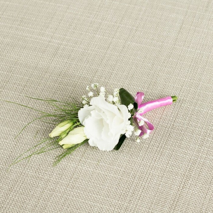 An elegant boutonniere lies at a slight diagonal across a neutral, light beige background that looks like natural linen or a similar woven fabric, its subtle texture adding warmth and softness to the composition. At the centre of the arrangement is a fully opened white lisianthus flower, its many ruffled petals forming a rounded bloom that appears almost weightless, the edges gently curling and catching the light. Just to the left and a touch lower, two slender lisianthus buds nestle close to the main flower, their creamy white tips fading into pale green stems, capturing that quiet moment just before they unfold. Threaded through the arrangement are delicate clusters of tiny white baby's breath, whose airy, cloud-like blossoms introduce a fine, romantic detail often favoured by couples marrying in churches and venues around South Croydon. Behind and around the flowers, slender, grass-like leaves fan out gracefully, extending towards the bottom left corner of the image, giving the boutonniere a flowing, organic line that feels like a small echo of the greenery in nearby Lloyd Park. A single, broader dark green leaf is tucked behind the main bloom, deepening the contrast against the white petals and helping the central flower stand out. At the base, the exposed stems are neatly gathered and wrapped with a vivid fuchsia satin ribbon, tied into a small, pert bow. This vibrant splash of pink introduces a contemporary, joyful accent against the more restrained palette of whites and greens. The photograph's gentle, even lighting highlights every petal edge and fibre of fabric, making the boutonniere appear fresh, crisp, and ready to be pinned to a groom's or groomsman's lapel for a South Croydon celebration. Crafted with precision and a florist's eye for balance, it evokes a sense of understated luxury and heartfelt occasion.