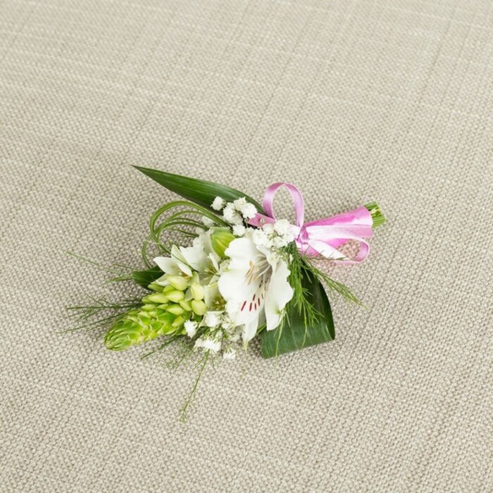 An elegant white and green boutonniere is carefully arranged on a light beige, slightly textured fabric background, reminiscent of natural linen set out on a dressing table in a South Croydon townhouse. The arrangement is compact but intricate, designed with the precision of an experienced florist. At its centre, a pristine white alstroemeria, also known as a Peruvian lily, opens fully, its petals broad and silky with a very gentle ruffle along the edges. Near the throat, fine reddish-brown streaks and speckles create delicate markings that draw the eye inward to the stamens, giving the bloom character and depth. Flanking the flower to the left is a tapering cluster of vibrant lime green buds, likely an Ornithogalum spike, whose tightly nested, scale-like buds lend a sculptural, almost geometric texture. Interwoven through the focal blooms are soft clouds of white baby's breath, tiny clustered blossoms that bring a sense of lightness and air, like a faint veil of mist. The entire piece is framed by lush greenery: dark green, pointed leaves provide a firm, supportive base, while long, fine strands of grass-like foliage curve gently around the flowers, adding movement that echoes the natural lines you might see in the planting around local parks such as Lloyd Park or along back gardens near Selsdon Road. On the right side, a neat bow crafted from glossy pink satin ribbon secures the stems; its smooth sheen and playful colour add a touch of celebration and romance without overpowering the classical palette. The light across the image is even and soft, revealing each texture-the velvety petals, crisp foliage, and silky ribbon-with clarity. The boutonniere feels fresh, refined, and ready to be pinned to a lapel for a wedding, prom, or formal gathering in South Croydon, a small yet expressive floral accent that speaks of care, elegance, and quiet joy.