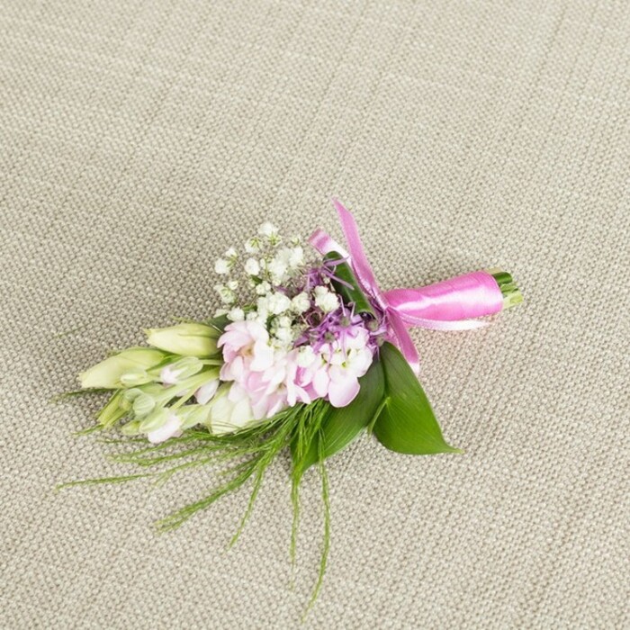 A romantic boutonniere, which could also serve as a small corsage, is carefully placed on a neutral, light beige linen backdrop, the fine weave of the fabric just visible and lending a soft, natural texture. The flowers themselves form a gentle oval, about the length of a hand, with the main cluster composed of ruffled, soft pink blooms resembling miniature stocks or garden-style spray flowers. Their petals overlap in layered waves, transitioning from a deeper rose-pink in the centre to a delicate, almost powdery blush at the edges, creating a sense of depth and softness. Interlaced with these are tiny sprays of white baby's breath, each stem branching into minuscule rounded blossoms that look like flecks of foam, bringing lightness and a slightly cloud-like effect. Small clusters of lavender or dusky purple florets peek out between the pink and white, giving just a hint of contrast, like distant heather on a hillside. To one side, several smooth, slender buds in creamy white and pale green stretch outward, still closed and elegant, reminiscent of freesias or similar elongated blooms, suggesting freshness and new beginnings. The entire composition is cradled in a bed of broad, rich green leaves, whose glossy surfaces catch the light, while finer, filament-like greenery and ferny sprigs thread around the edges to soften the outline. The stems at the base are firmly bound and finished with a bright fuchsia satin ribbon, wrapped neatly and tied into a symmetrical, tidy bow whose short tails curl slightly on the linen. The piece feels handcrafted and personal, like something prepared by a local South Croydon florist for a wedding near Croham Hurst Woods or a prom gathering off Pampisford Road, adding a polished yet heartfelt touch to formal attire.