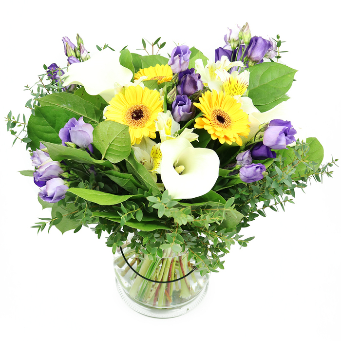 An opulent mixed bouquet in a clear glass cylindrical vase stands out against a bright white background. The top view reveals how the yellow gerberas anchor the arrangement, their large disc blooms radiating cheer with dark centers. White calla lilies add an elegant vertical note, while clusters of lavender lisianthus provide a soft, ruffled texture. Interspersed smaller white blooms, possibly alstroemeria, with gentle yellow markings, add delicate specks of light throughout.
Behind them, a lush tapestry of greens-glossy broad leaves, wispy fronds, and airy eucalyptus-gives depth and structure, while the transparent vase reveals neatly trimmed stems floating in fresh water. The bouquet exudes vitality, happiness, and timeless sophistication, ready for celebrations, gifts, or home decor. It speaks to the South Croydon customer seeking a refined, contemporary bouquet with a sunny twist, available for same-day delivery across Croydon and nearby towns.