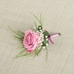 A single blush pink rose boutonniere lies carefully arranged on a softly textured beige linen surface, the kind of natural fabric you might find on a wedding table in a South Croydon reception venue. The image is taken from a slightly elevated, almost eye-level angle, so you can appreciate the depth of the rose as its velvety petals spiral outward. At the centre, the petals are a gentle pastel pink, gradually deepening to a more vivid rosy hue along the softly curled edges, suggesting a bloom cut at just the right moment of freshness. Clusters of tiny, cloud-like white baby's breath are tucked around the base of the rose, their rounded, pearl-sized blossoms creating a delicate halo and soft contrast against the pink. Slender, grass-like strands of light green foliage fan out from behind, wispy and relaxed, while a broader, darker green leaf is neatly folded to cradle the stems, giving the design structure and polish. The stems themselves are bound tightly with a smooth satin ribbon in harmonising tones of lavender-pink and deeper magenta, tied in a compact knot that sits neatly against the linen. A small silver-toned pin is visible near the base, ready to secure the piece to a lapel. The lighting is gentle and even, casting light shadows that emphasise the boutonniere's three-dimensional form without overwhelming it. The overall mood is romantic and composed, ideal for a groom or prom guest stepping out in South Croydon, perhaps near Lloyd Park or along Brighton Road, where a simple yet elegant floral accent can quietly express affection and celebration on an important day.