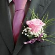 There's a genuine poetry in this South Croydon buttonhole, where every detail has been thoughtfully chosen to echo the spirit of meaningful gatherings. Nestled against a charcoal lapel, a softly blushed pink rose sits at the heart of the arrangement, its velvet petals opening just enough to suggest anticipation and tenderness-much like the mood at a celebration along Addiscombe Road or a wedding at local venues. The central rose is haloed by filaments of white baby's breath, delicate and cloud-like, lending a sense of serenity that's both modern and timeless. Wispy green leaves and deeper foliage arch supportively behind, introducing a natural energy reminiscent of the nearby green corridors. A gleaming satin ribbon in soft lavender gently secures the stems, its luster mirroring the muted radiance of the rose and complementing the bold fuchsia tie above a subtly latticed shirt. Framed by the rich hue and crisp tailoring of the jacket, the composition shines under soft daylight, capturing the essence of hope, unity, and celebration. To create such a boutonniere as a local florist is to infuse it with the community's warmth-a fragrant, beautiful keepsake meant to enhance milestones with a touch of South Croydon's unique charm.