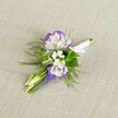 A finely detailed boutonniere is laid gently on a softly textured light beige linen background, the neutral cloth catching a calm, diffused light that lets every petal and leaf stand out. At the centre, two open lisianthus blooms glow in creamy white, their ruffled petals traced with a vivid band of deep purple that curls delicately at the edges, like ink brushed along a feathered edge. Tucked close to them, dainty clusters of tiny white filler flowers, much like baby's breath, create a cloudlike softness against the richer tones. Rising vertically from the heart of the design, a slender spike of grape hyacinth, or muscari, shows off tightly packed, bell-shaped florets in a strong, inky purple-blue, adding both height and texture. Around and beneath the blossoms, fine, feathery greenery-similar to asparagus fern-fans out in airy sprays, mingling with a few slightly broader green leaves to ground the composition and bring a sense of fresh growth. Two pale green, unopened buds sit alongside the open lisianthus, their smooth surfaces suggesting flowers that will unfurl later, echoing the quiet promise of a wedding morning. The visible stems are neatly aligned, expertly bound and wrapped in a crisp band of white satin ribbon, finished with a small, tidy bow that lies flat and ready for pinning to a lapel. The overall mood is polished yet romantic, the kind of handcrafted buttonhole you might see on a groom or prom guest stepping out onto a South Croydon side street near Lloyd Park, carefully created to add a subtle touch of floral elegance to formal attire without overwhelming it.