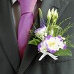 A close-up wedding portrait focuses on the upper torso of a groom dressed in a dark charcoal grey suit, standing as if just outside a South Croydon church on a bright afternoon. The suit jacket is cleanly tailored with a sharp lapel, layered over a light grey checkered dress shirt that adds a subtle, refined pattern. At the centre of the image, a lustrous magenta tie is perfectly knotted, its silk surface catching the light with a soft sheen that echoes the joyous mood of a wedding morning near Lloyd Park. Pinned to the left lapel is an exquisitely crafted boutonniere: two open white lisianthus blooms edged with deep violet ruffles and glowing yellow centres, their petals appearing almost porcelain-smooth. Around them, several slim, unopened white buds suggest new beginnings, while a single slender spike of deep indigo-purple veronica rises gracefully, giving gentle height and structure. Tiny clouds of white filler flowers, similar to baby's breath, create a delicate halo, threaded through with airy sprigs of feathery greenery and broader emerald leaves that curve protectively around the blooms. Fine loops of decorative green grass or wire add a modern, sculpted twist, like a designer detail you might notice at a stylish South Croydon reception venue. At the base, a small, pristine white satin ribbon is tied into a neat bow, its soft, light-catching folds grounding the design in classic tradition. The overall effect is one of quiet sophistication and heartfelt celebration, as though the wearer is ready to step into a day of promises, photos, and cherished memories shared with friends and family close to home.