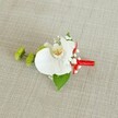 A refined wedding boutonniere rests on a softly textured, light beige linen background, its neutral tone reminiscent of the stone facades and calm streets of South Croydon on a quiet morning. At the heart of this floral pin sits a single white phalaenopsis orchid, its broad, velvety petals opening with understated grace. The orchid's centre glows with soft yellow and touches of reddish-orange or light brown markings, like tiny brushstrokes that draw the eye into its throat. Around it, clusters of delicate white baby's breath create a misty, cloud-like halo, their tiny blossoms adding airiness and a sense of gentle movement. To the left, two round lime-green chrysanthemum buds, packed with fine, spiky petals, introduce a modern burst of fresh colour, echoing the vibrant greens seen in nearby Lloyd Park or along the tree-lined roads by South Croydon station. Beneath the flowers, a rich, deep green leaf supports the arrangement, with a smaller, darker leaf peeking near the base, adding depth and structure. The stems are neatly wrapped in a vivid crimson red satin ribbon, the smooth fabric tightly coiled and finished with a small, tidy knot that lends a touch of festive formality, as if ready to be pinned on a groom just before a ceremony at a local church. The weave of the linen background is clearly visible, lending a natural, slightly rustic character that contrasts beautifully with the refined flowers. The overall mood is one of quiet elegance and careful craftsmanship, the sort of bespoke detail a South Croydon florist might prepare for a groom, groomsman, or prom guest wanting something both classic and contemporary, fresh and memorable.
