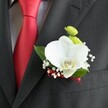 A close-up photograph focuses tightly on a groom's lapel, where a refined wedding boutonniere rests against the deep charcoal weave of a tailored suit jacket, the fabric showing a subtle, elegant texture in the light. At the heart of the arrangement is a large white Phalaenopsis orchid, its broad, velvety petals curving outward like soft wings and framing a delicate yellow-and-white lip that glows gently at the centre. Nestled just above the orchid, two small, round lime-green chrysanthemum-style blooms add a bright, playful contrast, their tiny petals forming neat, cushion-like domes of colour. Around these focal flowers, clusters of airy white baby's breath are tucked in, each minuscule blossom forming a misty halo that softens the composition and gives the design a romantic, almost cloudlike feel. A few glossy, rich green leaves peek from behind, grounding the floral work with a natural depth and freshness. The base of the boutonniere is wrapped in a slim deep red satin ribbon that echoes the bold red of the groom's tie, which runs sharply down the centre of a light grey and white small-checked dress shirt to the left of the frame. The interplay of red satin, white orchid, and green accents creates a harmony that feels contemporary yet timeless, perfectly suited to a South Croydon wedding, perhaps in a church near Croham Road or a reception by Lloyd Park. The lighting is soft and flattering, catching the gentle sheen of petals and fabric, and every detail speaks of quiet luxury, careful craftsmanship, and the joyful anticipation of a day devoted to love and celebration.