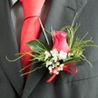 The image offers a close, intimate view of a lovingly designed boutonniere, pinned to the lapel of a dark charcoal grey suit that could easily belong to a groom heading to a South Croydon church or registry office near Park Hill Gardens. Center stage is a cerise-pink rosebud, partly opened so that its velvety petals fold around each other in a soft spiral, full of colour and quiet intensity. Encircling the rose is a gentle cloud of white baby's breath, each tiny flower precise and airy, creating a soft halo that contrasts beautifully with the bold central bloom. A combination of fresh greenery supports the composition: slender, feathery stems adding lightness, and fuller, glossy leaves bringing a sense of structure and depth, as though each piece was carefully chosen on the florist's workbench earlier that morning. At the base, a vivid scarlet satin bow is tied neatly, its bright, smooth ribbon catching the light and echoing the rich red of the silk necktie tucked beneath the lapel. The suit's fabric appears to be a fine wool blend, with a subtle, tailored texture, while a grey dress shirt with a faint pattern provides a sophisticated backdrop. Gentle, diffused lighting accentuates the contours of the rose, the crisp white gypsophila, and the sheen of the ribbon without overwhelming the scene. The result is a refined, romantic buttonhole that feels personal and intentional, perfectly suited for a South Croydon wedding day filled with anticipation, heartfelt vows, and the quiet pride of dressing for one of life's most meaningful moments.