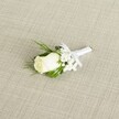 A finely crafted wedding boutonniere is displayed diagonally across a softly woven, light beige fabric that resembles natural linen, its gentle crosshatch texture giving the image an understated, elegant backdrop. At the heart of the design sits a single creamy white rosebud, its velvety petals still mostly furled, with just the faintest suggestion of unfurling at the center, like a quiet promise at the start of the day. The rose's base shows hints of fresh green sepals, emphasising its youth and freshness, as if it has just been prepared in a South Croydon workshop before being delivered to a groom getting ready near Croham Road or Lloyd Park. Surrounding the rose are airy clusters of white baby's breath, each tiny bloom forming a cloud-like halo that softens the outline of the central flower and adds an ethereal lightness. Slender, deep green leaves and a few delicate, fern-like sprigs extend from behind the rose, their smooth and feathery textures creating subtle layers of depth and movement. The stems are neatly bound with crisp white satin ribbon, wrapped tightly and finished in a small, carefully tied bow positioned just beneath the bloom, ready to sit flat against a lapel. Soft, diffused lighting glides over the composition, catching the gentle sheen of the ribbon, the satiny surface of the rose petals, and the matte weave of the fabric below. The overall feeling is one of classic refinement and quiet romance, the kind of boutonniere chosen for a traditional church ceremony in South Croydon or a simple registry office wedding, symbolising purity, new beginnings, and the thoughtful care that goes into every detail of a special day's attire.