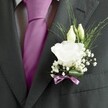 A close-up photograph of a groom's charcoal grey suit in soft, natural light, focusing tightly on the left lapel where a refined white lisianthus buttonhole has been pinned. The wool suit jacket has a subtle woven texture and a sharply pressed lapel, framing a boldly knotted magenta silk tie that catches the eye against a patterned white and grey dress shirt. Nestled at the heart of the image is the boutonnière itself: a fully opened white lisianthus bloom with gently ruffled petals, full and velvety, surrounded by two slender ivory buds that look ready to unfurl. Wisps of fine greenery arc outwards, delicate and linear, while clusters of tiny white gypsophila create a cloud-like halo around the central flower. The base of the arrangement is wrapped neatly in a narrow mauve ribbon, tied with care so the colour echoes the tie and adds a quiet touch of romance. The flowers appear freshly arranged, as if prepared that morning in a South Croydon florist's studio for a wedding at a nearby church or registry office. The overall mood is one of calm anticipation and elegant celebration: the contrast between the dark suit and the pale blooms suggests a moment just before the ceremony begins, perhaps on a leafy street near Lloyd Park or strolling up from South Croydon station, where the wearer stands poised, dressed with subtlety and intention for a day filled with vows, photographs, and heartfelt toasts.