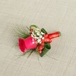 A meticulously crafted boutonniere rests on softly textured, light beige linen, the neutral fabric echoing the understated elegance of a South Croydon wedding morning. At its heart sits a vivid fuchsia-pink rosebud, petals velvety and tightly layered, just beginning to unfurl in a perfect spiral that suggests freshness and anticipation. Nestled around the rose are delicate clouds of tiny white baby's breath blossoms, their airy clusters softening the bold colour and adding a touch of classic romance. Threaded between these miniature blooms are bright red hypericum berries, glossy and plump, catching the light with a gentle sheen and introducing a festive, almost jewel-like accent. Encircling the composition, deep green folded leaves create a structured collar, while finer, wispy, fern-like greenery extends outward, giving the design a sense of movement and garden-grown naturalness. The stems are neatly bound with a narrow, bright red satin ribbon, wrapped smoothly and tied into a small, precise knot that feels both secure and luxurious to the touch. Gentle, diffused lighting from above casts soft shadows across the woven fabric, highlighting every curve of petal and leaf. The overall impression is one of intimacy and celebration: a compact floral piece designed to be pinned to a lapel for a wedding, prom, or black-tie event in and around South Croydon, perhaps worn on the walk from a ceremony near Lloyd Park to an evening reception. It feels like the kind of handcrafted detail chosen with care, a quiet statement of joy that complements tailored suits and cherished local celebrations alike.