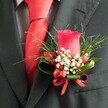 A close-up, high-angle photograph reveals a meticulously crafted boutonniere pinned to the left lapel of a dark charcoal suit jacket, the kind you might see on a groom stepping out of a car near South Croydon station on a bright wedding morning. At the centre sits a single, perfectly formed deep red rose bud, its velvety petals tightly layered and upright, glowing like a ruby against the dark wool of the jacket. Encircling the rose, a halo of delicate white filler flowers, much like tiny sprays of baby's breath, adds a soft, cloud-like contrast that lightens the composition. Nestled between the blossoms are several small, glossy red berries, oval and plump, echoing the rose's rich hue and adding a touch of texture and seasonal charm. Lush greenery anchors the design: slender, grass-like fronds fan out in gentle arcs, while broader, darker green leaves curve protectively around the base, giving the boutonniere structure and depth. A narrow red satin ribbon is wrapped neatly around the stems and tied into a precise little bow, its sheen catching the indoor light and tying together the red elements with quiet sophistication. Just beneath the lapel, a vivid red tie in a smooth, likely silk or satin fabric is visible, its knot crisply executed and perfectly centred, mirroring the elegance of formal events held around Lloyd Park or wedding receptions in nearby venues. Behind the tie, a subtly patterned dress shirt in light grey and white-perhaps a fine check or houndstooth-adds understated texture, completing an ensemble that feels polished, romantic, and thoughtfully styled for a special occasion in South Croydon.