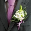 A close-up, high-angle photograph shows the upper torso of a man dressed for a formal occasion in South Croydon, his dark charcoal grey suit jacket filling most of the frame. The fabric looks smooth and finely tailored, with a crisp lapel line that draws the eye towards a pale, grid-patterned dress shirt and a perfectly knotted lavender tie running straight down the centre of his chest. Pinned carefully to the left lapel is a handcrafted boutonniere, its fresh flowers standing out vividly against the deep charcoal cloth. At the top, a slim, light green spiky bloom rises upright, like a hyacinth or unopened Muscari, giving the arrangement height and a gentle sense of anticipation. Below it, open white alstroemeria blossoms unfurl, each petal marked with delicate burgundy streaks and tiny amber stamens that catch the soft natural light, much like the light that filters through gardens off Brighton Road or near Purley Oaks. Wisps of white baby's breath weave between the larger blooms, their tiny clustered florets creating a misty, romantic halo. Surrounding everything is a mix of lush greenery: slender, grass-like blades curve and twist, while broader, darker leaves provide a grounding backdrop. At the base, a satin lilac ribbon, perfectly matched to the tie, is knotted into a neat bow that hugs the stems and foliage together. The overall mood is one of calm elegance and quiet excitement, the kind of refined detail you might notice just before stepping out for a wedding ceremony in a South Croydon church, registry office, or intimate garden venue, the cool petals and fresh green scent offering a moment of stillness before the celebration begins.