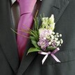 A close-up wedding portrait focuses tightly on the upper chest of a man in a sharply tailored charcoal suit, the kind you might see on a groom stepping out of a car near South Croydon station. The jacket's dark, subtly textured lapel frames a crisp, pale dress shirt with a faint check pattern, just visible at the edges. At the centre runs a bold magenta silk tie, neatly knotted and gently glowing where the light catches its smooth, lustrous surface, adding a confident splash of colour. Pinned precisely to the left lapel is an intricate boutonnière, clearly crafted with the care of an experienced florist. A few slender, unopened cream buds rise at the top, echoing the soft elegance of snapdragons. Beneath them sit clusters of pastel pink and light lavender blooms, reminiscent of sweet peas or stock, cupped by tiny clouds of white gypsophila that bring an airy, dewy feel, like a morning walk through nearby Lloyd Park. Deeper purple florets are tucked in for contrast, while glossy dark green leaves create a strong, structured base. Fine, feathery sprigs of brighter green foliage arc outwards, giving movement and lift. Around the stems, a delicate lavender satin ribbon is tied into a neat bow, its sheen subtly echoing the tie's richness without competing with it. The softly diffused daylight, perhaps from a townhouse window off Brighton Road, bathes everything in a warm, flattering glow. Together, the flowers, fabrics, and colours create a poised yet playful look, perfect for a groom or honoured guest at a South Croydon wedding, capturing both celebration and quiet anticipation in a single, refined detail.